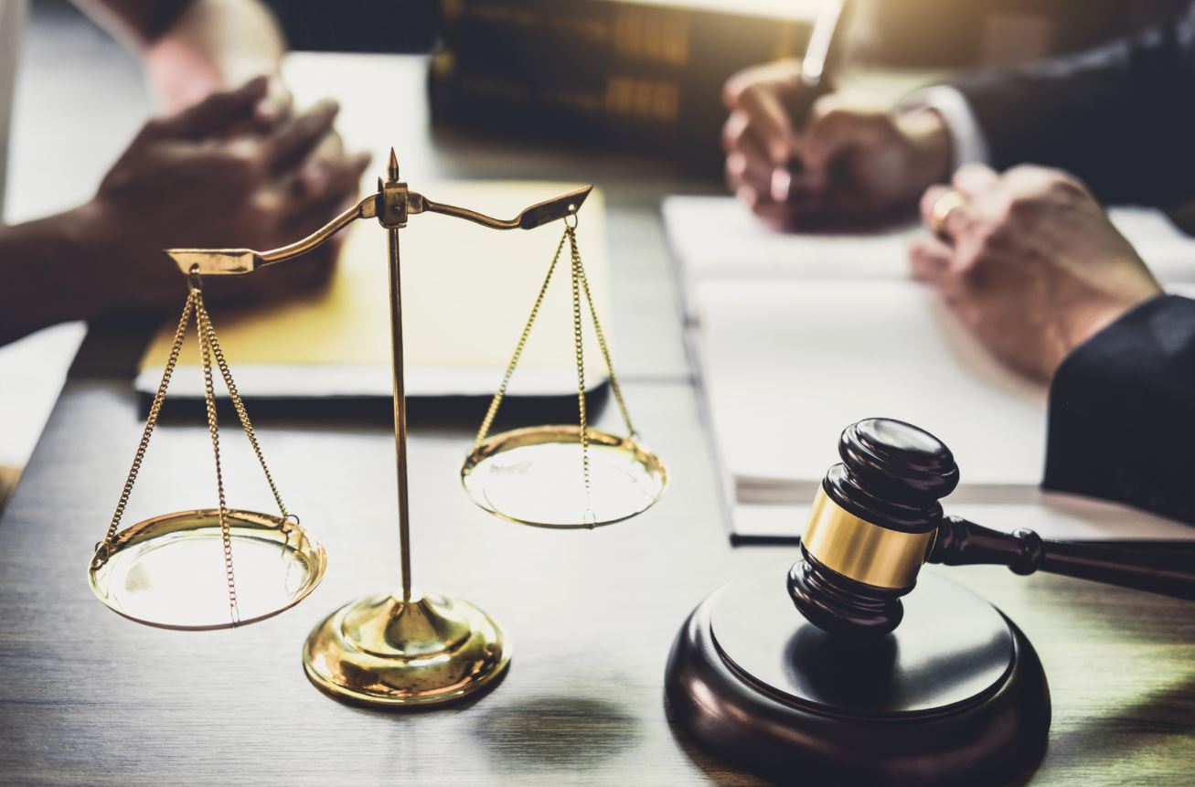 A close-up of a legal setting featuring a scale of justice and a gavel, with hands engaged in discussion and note-taking.